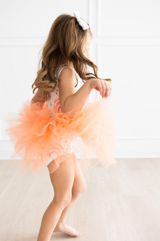 Child in an orange tutu sitting on a light-colored floor with a white background