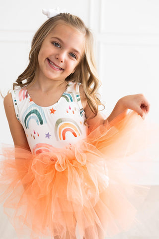 Young girl wearing a leotard with rainbow designs and an orange tutu.
