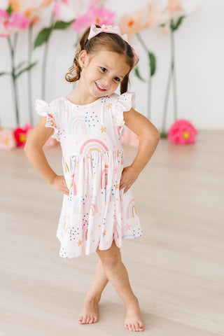 Young girl wearing a light pink dress with colorful designs, standing in front of flowers.