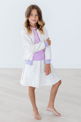 Young girl wearing a white dress with a purple top and white cardigan on a light background