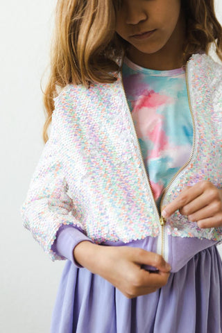 Child wearing a sequined jacket over a colorful shirt and purple skirt on a plain background