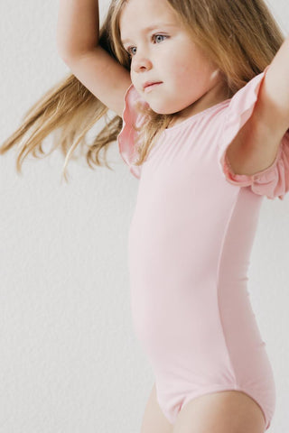 Young girl in a pink leotard with arms raised against a light background