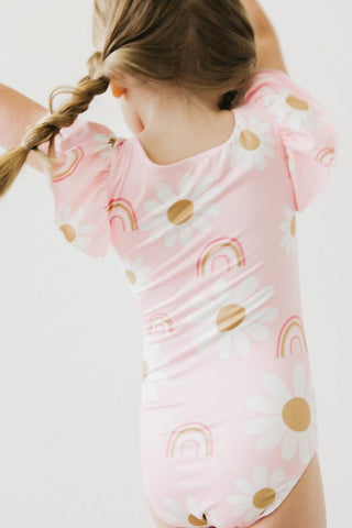 Child wearing a pink leotard with rainbow and cloud patterns on a white background